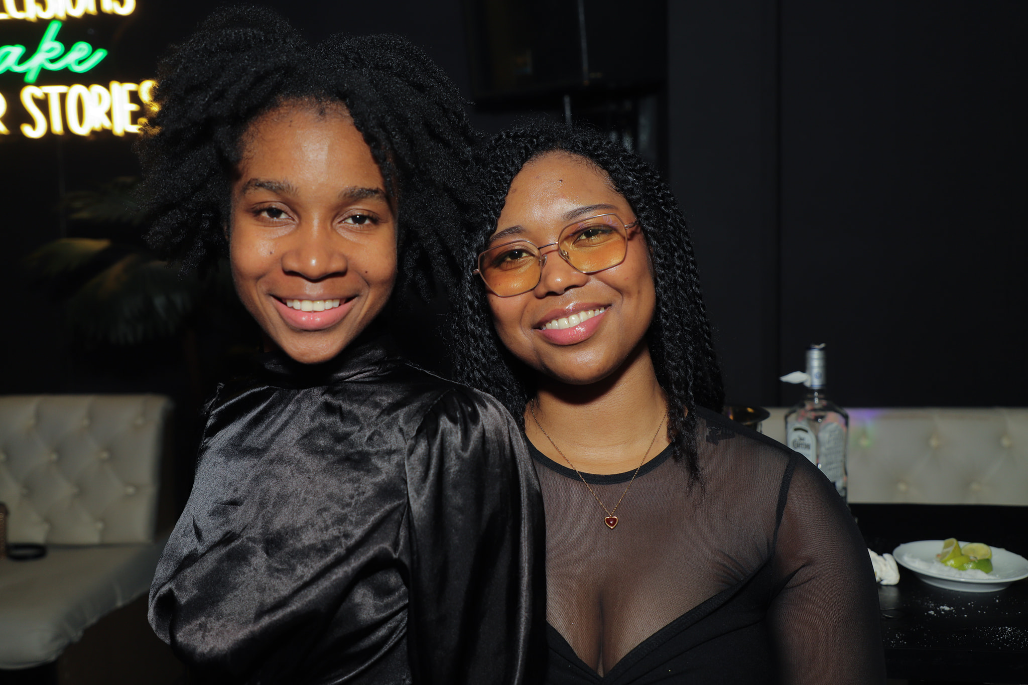 Two friends smiling in front of a neon sign that reads 'make better stories'.